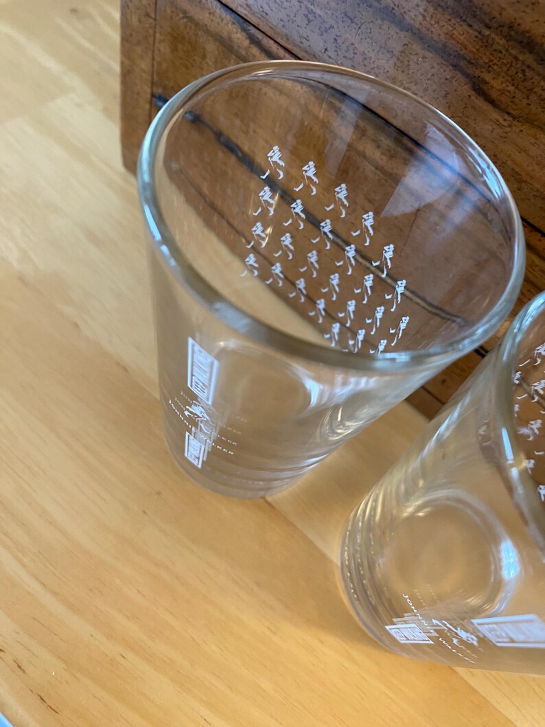 Op de afbeelding: Twee heldere glazen bekers met witte bedrukte ontwerpen. E&eacute;n beker heeft kleine figuren, de andere een gebouwontwerp. De glazen staan op een licht houten oppervlak.