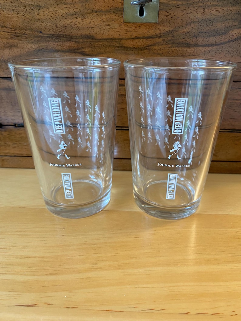 Op de afbeelding: Twee heldere glazen met de tekst "KEEP WALKING" en "JOHNNIE WALKER" in het wit gedrukt. De glazen hebben een ontwerp van een wandelende figuur en een menigte mensen. De glazen staan op een houten oppervlak.
