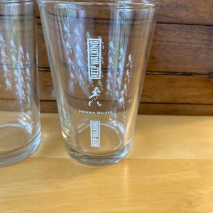 Op de afbeelding: Twee heldere drinkglazen met de tekst "KEEP WALKING" en het Johnnie Walker-logo. De glazen hebben een reeks kleine witte figuren die rond het glas lopen. De glazen staan op een lichtbruin houten oppervlak.