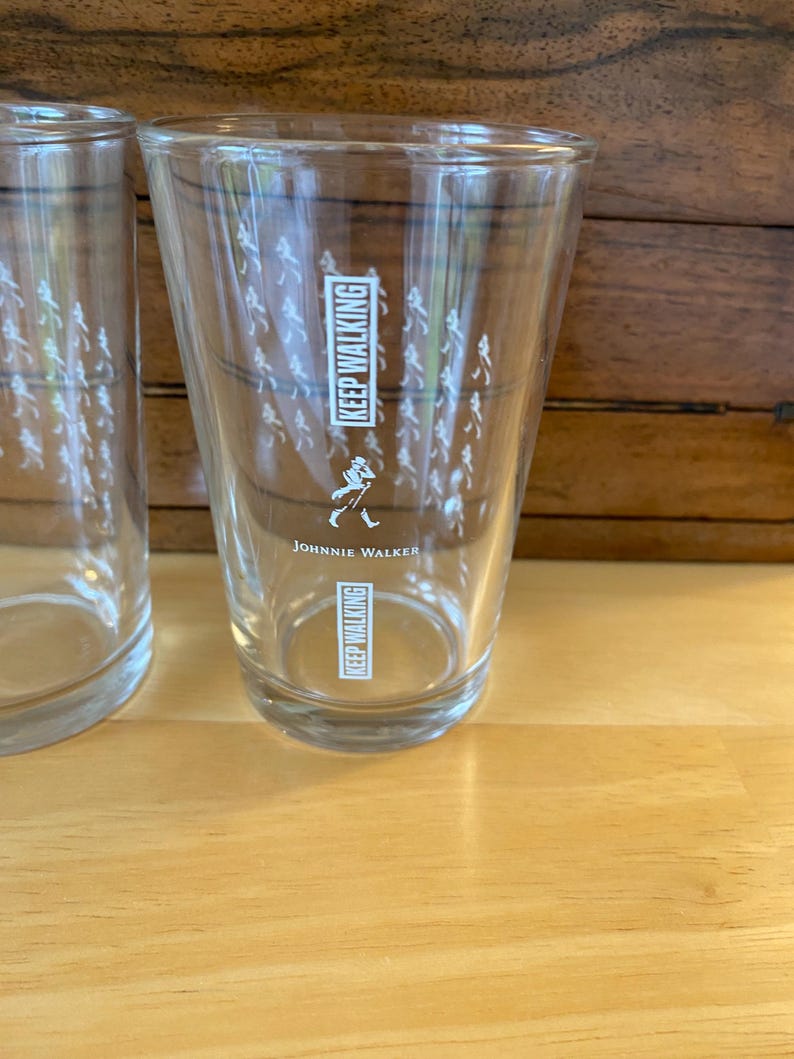 Op de afbeelding: Twee heldere drinkglazen met de tekst "KEEP WALKING" en het Johnnie Walker-logo. De glazen hebben een reeks kleine witte figuren die rond het glas lopen. De glazen staan op een lichtbruin houten oppervlak.