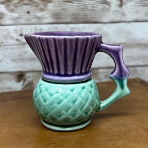 Könnte beinhalten: Kleine, dekorative Keramikkrug mit einzigartigem Design. Die Oberseite ist gerippt lila, die Mitte ist strukturiert grün und der Griff ist lila und grün. Der Krug steht auf einer braunen Oberfläche.