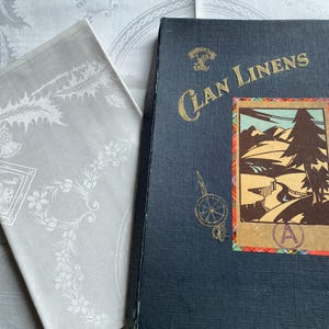 Op de afbeelding: Een gevouwen witte linnen servet met een bloemen- en hulstontwerp ligt naast een donkerblauw boek met de titel "Clan Linens". Het boek heeft een kleurrijke landschapsillustratie en gouden letters.