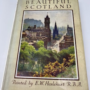 Puede incluir: Un libro vintage titulado "Beautiful Scotland" con una ilustración pintada de una ciudad en la portada. La ilustración representa una vista de Edimburgo, Escocia, con el icónico Monumento a Scott y el Castillo de Edimburgo en el fondo. El libro es de E.W. Haslehust R.B.A.