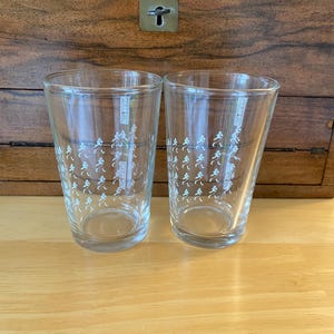 Op de afbeelding: Twee heldere glazen glazen met een herhaalde witte afbeelding van een rennende persoon. De glazen zijn cilindrisch met een licht uitlopende bovenkant. De achtergrond is een houten kast.
