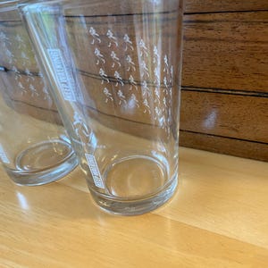 Op de afbeelding: Twee heldere drinkglazen met de woorden "KEEP WALKING" verticaal afgedrukt. De glazen hebben een patroon van kleine witte figuren die lopen. De glazen staan op een lichtbruin houten oppervlak.