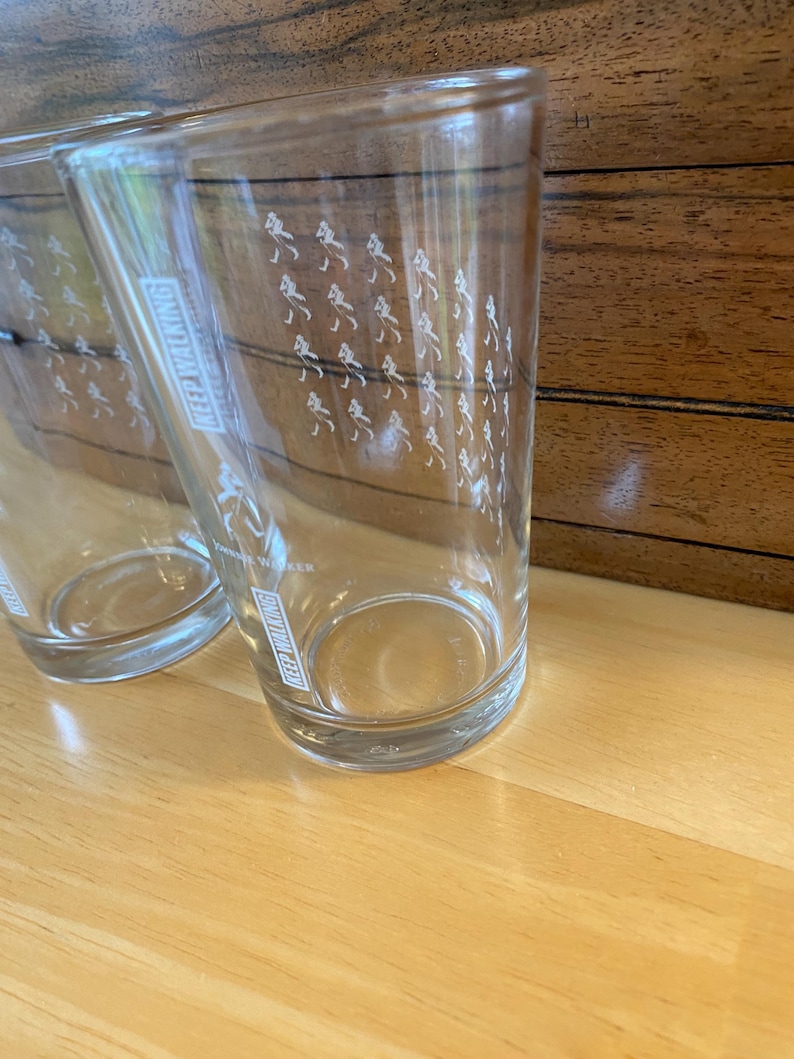 Op de afbeelding: Twee heldere drinkglazen met de woorden "KEEP WALKING" verticaal afgedrukt. De glazen hebben een patroon van kleine witte figuren die lopen. De glazen staan op een lichtbruin houten oppervlak.