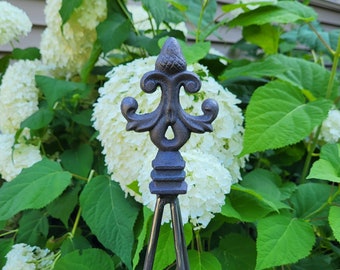 May include: A black metal garden stake with a decorative fleur-de-lis finial. The stake is in front of a bush of white hydrangeas.