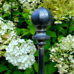 May include: A dark gray metal garden stake featuring a spherical finial top with a textured, hammered surface. The finial sits atop a decorative, tiered base that connects to a straight, slender metal rod extending downward from the center.