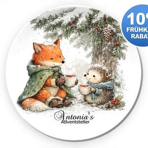 Könnte beinhalten: Ein runder weißer Teller mit einer Aquarellillustration eines orangefarbenen Fuchses im grünen Umhang und eines braunen Igels mit blauem Schal, die beide Tassen unter einem verschneiten Tannenbaum halten. Unten steht der Text Antonia's Adventsteller.