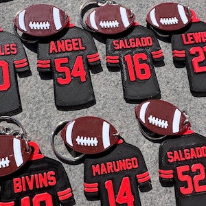 May include: A set of 10 black and red football jersey keyrings with a football charm. The jerseys have different names and numbers on them, including "Angel 54", "Salgado 16", "Lewis 2", "Bivins 10", and "Marungo 14".