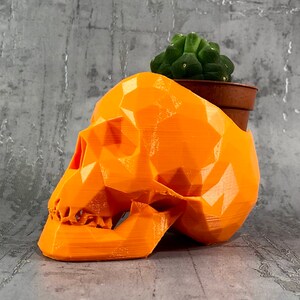Peut inclure: Planteur en forme de crâne géométrique orange avec une petite plante de cactus verte à l'intérieur. Le planteur est en plastique imprimé en 3D.