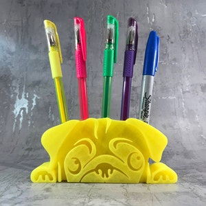 May include: A yellow pug-shaped desk organizer with five pens sticking out of the top. The pens are yellow, red, green, purple, and silver. The silver pen has the word "Sharpie" written on it.