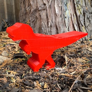 May include: A bright red plastic dinosaur pencil holder. The dinosaur is standing on a bed of brown mulch.