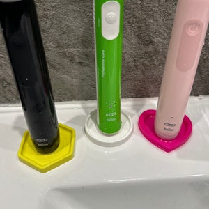 Könnte beinhalten: Drei elektrische Zahnbürsten mit unterschiedlich farbigen Griffen und Sockeln. Die Zahnbürsten sind schwarz, grün und rosa. Die Sockel sind gelb, weiß und rosa. Die Zahnbürsten sind mit "Oral-B" beschriftet.