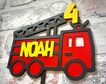 May include: Red and black fire truck cake topper with a yellow ladder, the name "NOAH" and the number "4" on top.