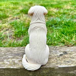 May include: A cream-colored dog figurine, viewed from the back, sits on a wooden surface against a backdrop of green grass. The dog's fur is detailed with textured lines, and its tail curls up. The figurine is a decorative item.