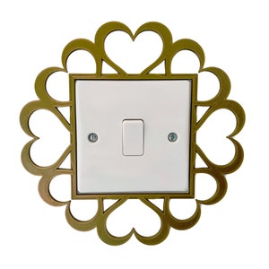 Puede incluir: Un interruptor de luz blanco con un marco decorativo dorado con un diseño de corazón. El marco está hecho de un material delgado e intrincado.