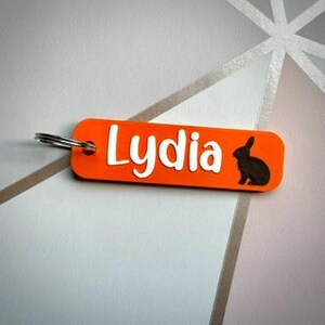 Puede incluir: Llavero rectangular naranja con el nombre "Lydia" en blanco y una silueta negra de conejo. El llavero tiene un llavero plateado.