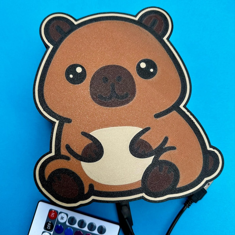 May include: A brown bear-shaped LED lamp with a black outline and a USB cable. The lamp has a light beige belly and a dark brown nose and ears. A remote control with buttons is visible at the bottom. The background is a bright blue.