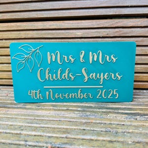 Op de afbeelding: Een turquoise rechthoekig bord met gouden letters en een bladerontwerp. De tekst luidt "Mrs & Mrs Childs-Sayers 4th November 2025". Het bord is waarschijnlijk voor een bruiloft of een speciale gebeurtenis.