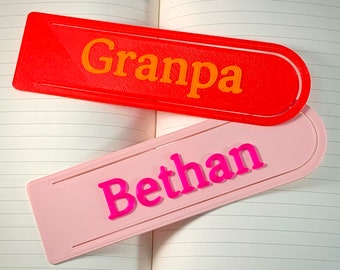 Könnte beinhalten: Zwei personalisierte Lesezeichen, eines rot mit dem Namen "Grandpa" in orangefarbenem Text und eines rosa mit dem Namen "Bethan" in rosa Text.