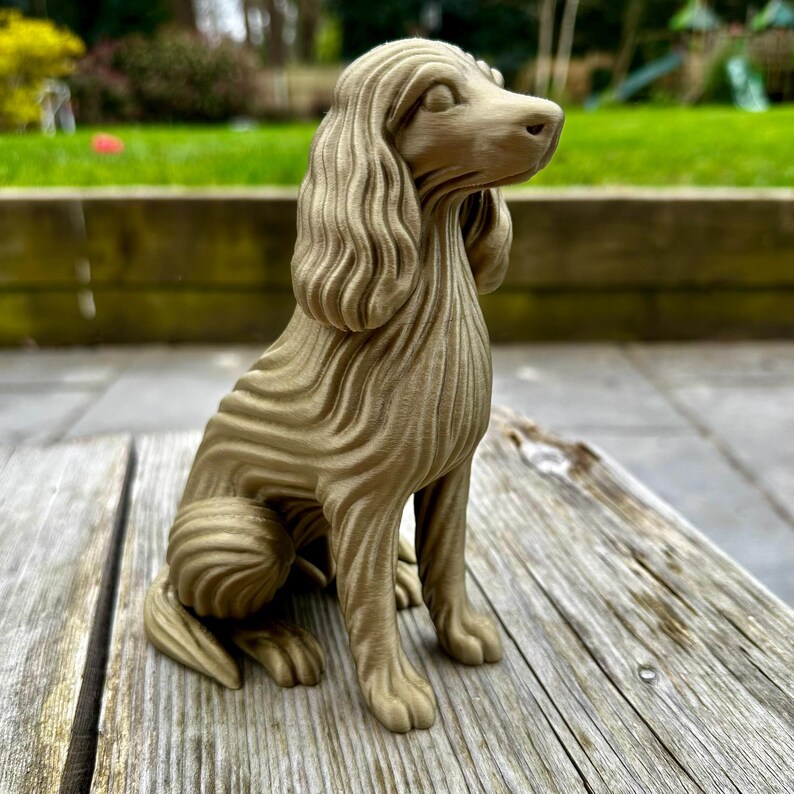 May include: A detailed, golden-colored dog figurine, sculpted with a unique ridged texture. The dog is in a seated position, with its head turned slightly to the right. The figurine is placed on a weathered wooden surface, with a blurred green background.
