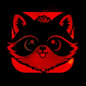 May include: A glowing red raccoon face design on a black background. The raccoon has large eyes, a small nose, and a smiling mouth with its tongue sticking out. The image is a square shape.