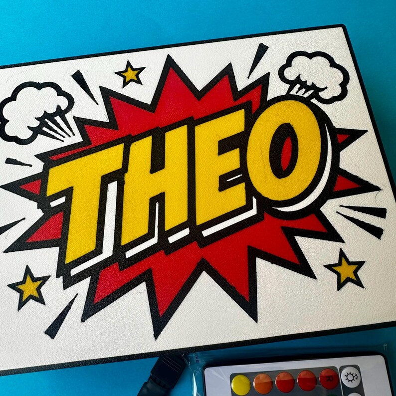 May include: A white sign featuring the name "THEO" in yellow, outlined in black, within a red burst design. The artwork includes star and cloud motifs. A remote control with coloured buttons is visible at the bottom.