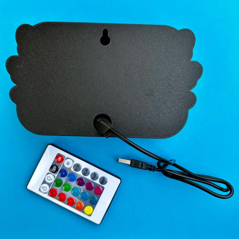 K&ouml;nnte beinhalten: Ein schwarzes, dekoratives Schild mit gewelltem Rand, einem USB-Kabel und einer Fernbedienung mit mehreren Farboptionen. Die Fernbedienung hat Tasten f&uuml;r verschiedene Farben und Einstellungen. Der Hintergrund ist einfarbig blau.