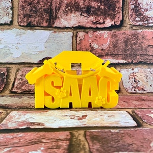 Peut inclure: Une maison pour chien en 3D imprimée en jaune avec le nom "ISAAC" écrit en grandes lettres. La maison a deux accents en forme d'os sur les côtés et des empreintes de pattes sur le devant.