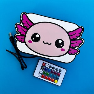 K&ouml;nnte beinhalten: Ein Nachtlicht in Axolotl-Form mit rosa K&ouml;rper, schwarzen Augen und lila Kiemen. Eine Fernbedienung mit Farboptionen und ein USB-Kabel sind enthalten. Der Hintergrund ist einfarbig blau.