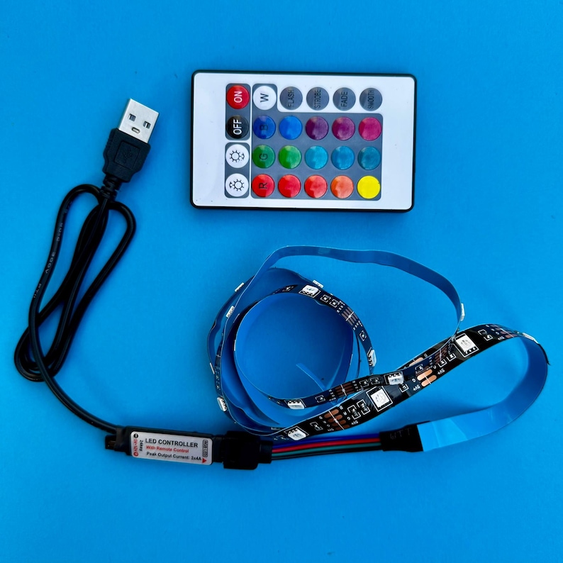May include: LED strip lights with a remote control and USB cable, displayed on a blue surface. The remote has buttons for color selection and light effects. The LED strip is black with small lights.