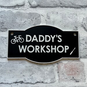 May include: Black and white metal sign with rounded edges that reads "Daddy's Workshop" with a bicycle and screwdriver icon.
