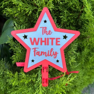 Puede incluir: Un letrero en forma de estrella roja y blanca con el texto "The WHITE Family" en una fuente de escritura. El letrero tiene un borde rojo, un centro azul claro y recortes de estrellas negras. Un clip y una cinta rojos est&aacute;n adjuntos en la parte inferior.