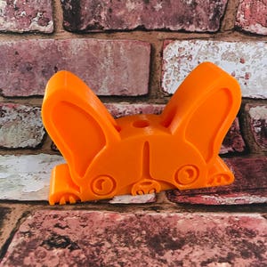 May include: Orange plastic figurine of a French bulldog with its head down and ears perked up. The figurine is a 3D printed model.