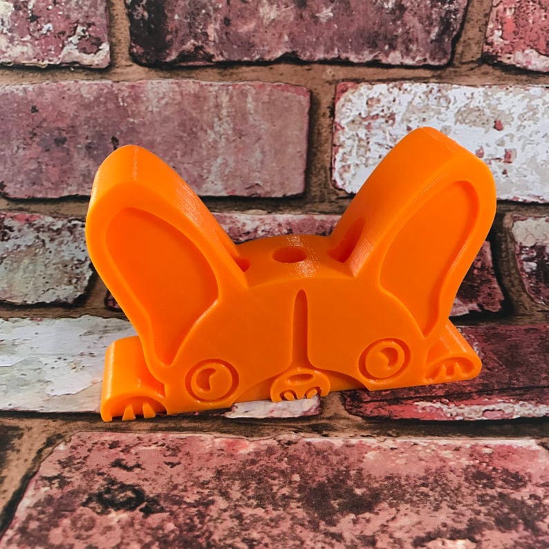 May include: Orange plastic figurine of a French bulldog with its head down and ears perked up. The figurine is a 3D printed model.