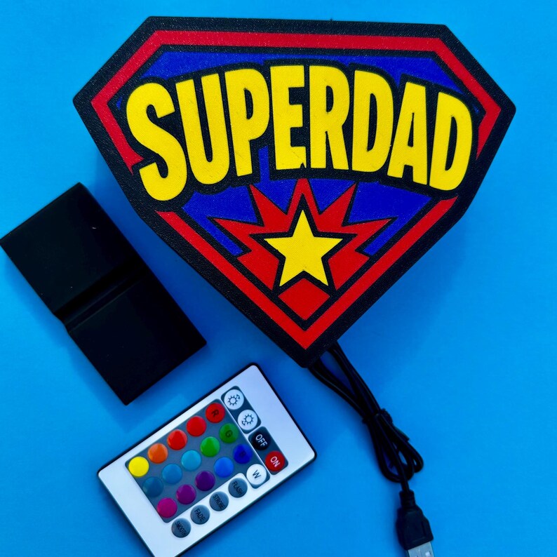 May include: A Superdad sign in a shield shape with the text "SUPERDAD" in yellow. The sign has a red, blue, and yellow color scheme. A remote control and a black USB cable are also visible.