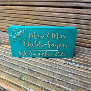Op de afbeelding: Een turquoise rechthoekig bord met gouden letters en een bladerontwerp. Op het bord staat "Mrs & Mrs Childs-Sayers 4th November 2025". Het bord is gemaakt van een stevig materiaal en staat op een houten oppervlak.