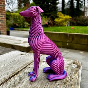 May include: A decorative sculpture of a dog in a seated position. The dog is a vibrant magenta color with blue accents, featuring a textured, wave-like pattern. The sculpture is placed on a weathered wooden surface.