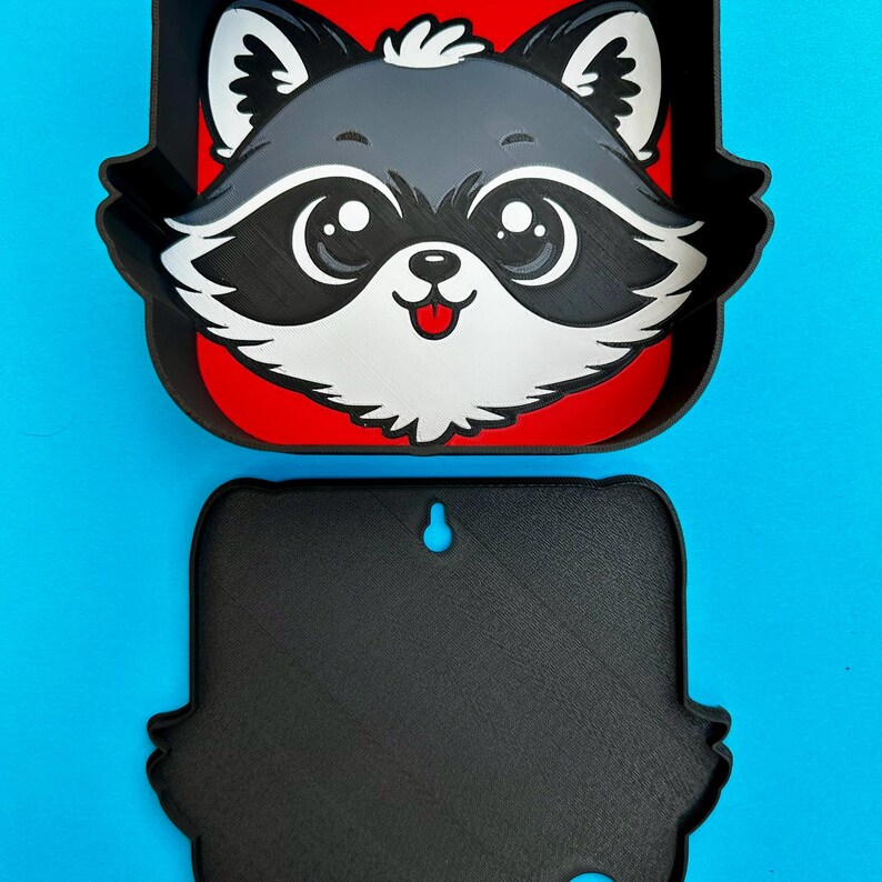 May include: A two-piece, black and red raccoon-themed wall decor. The top piece features a raccoon face with gray fur, black markings, and a red background. The bottom piece is a solid black, raccoon-shaped piece with a hole for hanging. The background is a solid blue.