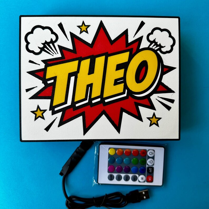 May include: A rectangular light-up sign in a comic book style, featuring the name "THEO" in yellow with a black outline, set against a red and white backdrop. Includes a remote control and USB cable.