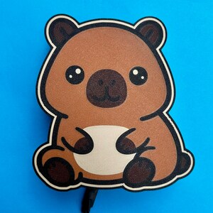 May include: A brown bear-shaped sticker with a white belly, black eyes, and a black nose. The sticker has a black border and is set against a bright blue background. The bear is in a sitting position.