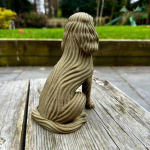 May include: A detailed, seated dog sculpture with a textured, flowing design. The sculpture is a muted olive green color and sits on a weathered wooden surface. The dog's form is stylized, with a focus on the texture of its fur.