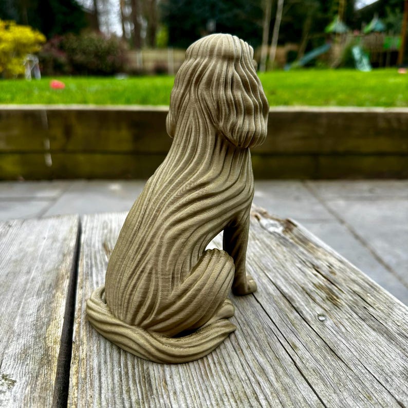 May include: A detailed, seated dog sculpture with a textured, flowing design. The sculpture is a muted olive green color and sits on a weathered wooden surface. The dog's form is stylized, with a focus on the texture of its fur.