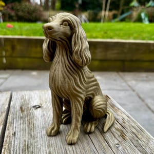 May include: A detailed, golden-brown dog figurine, seated with its head tilted upwards. The sculpture features textured fur and a long, flowing ear. The dog is positioned on a weathered wooden surface, with a blurred green background.