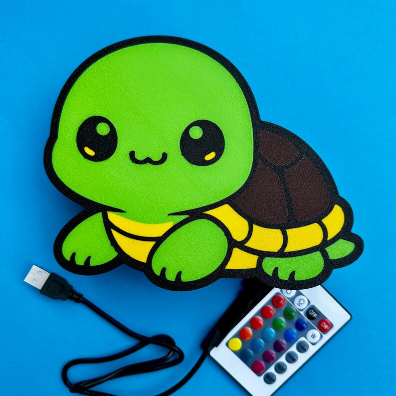 K&ouml;nnte beinhalten: Eine gr&uuml;ne, schildkr&ouml;tenf&ouml;rmige LED-Lampe mit braunem Panzer und gelben Akzenten. Sie verf&uuml;gt &uuml;ber ein USB-Kabel und eine Fernbedienung mit Farbauswahl. Der Hintergrund ist einfarbig blau.