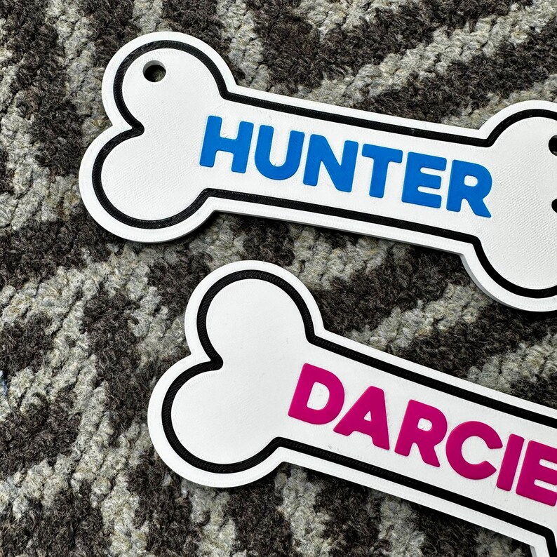 Puede incluir: Dos etiquetas para mascotas con forma de hueso. Una etiqueta tiene el nombre "HUNTER" en letras azules. La otra etiqueta tiene el nombre "DARCIE" en letras rosas. Ambas etiquetas son blancas con un contorno negro y un agujero para sujetar.
