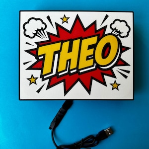 May include: A white rectangular sign featuring the name "THEO" in yellow, with a black outline, set against a red burst background. The design incorporates comic book style elements such as stars and explosion clouds. A black USB cable is attached.