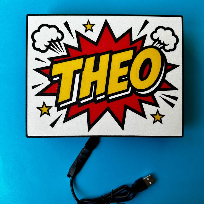 May include: A white rectangular sign featuring the name "THEO" in yellow, with a black outline, set against a red burst background. The design incorporates comic book style elements such as stars and explosion clouds. A black USB cable is attached.