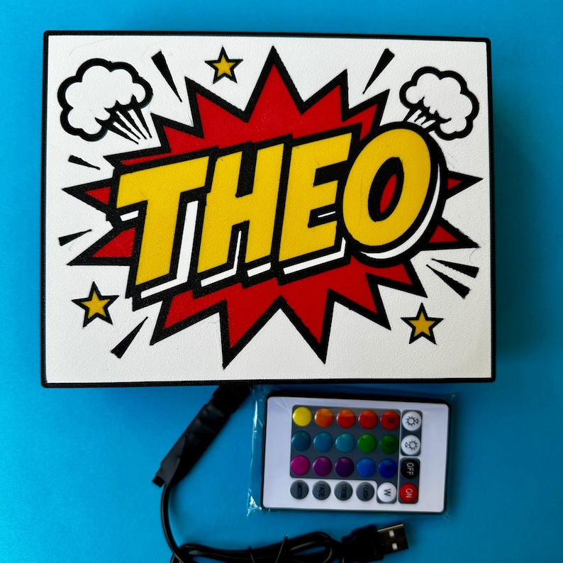 May include: A white sign in a comic book style, featuring the name "THEO" in yellow with a black outline, set against a red burst. The design includes white cloud and star accents. A remote control and USB cable are visible.